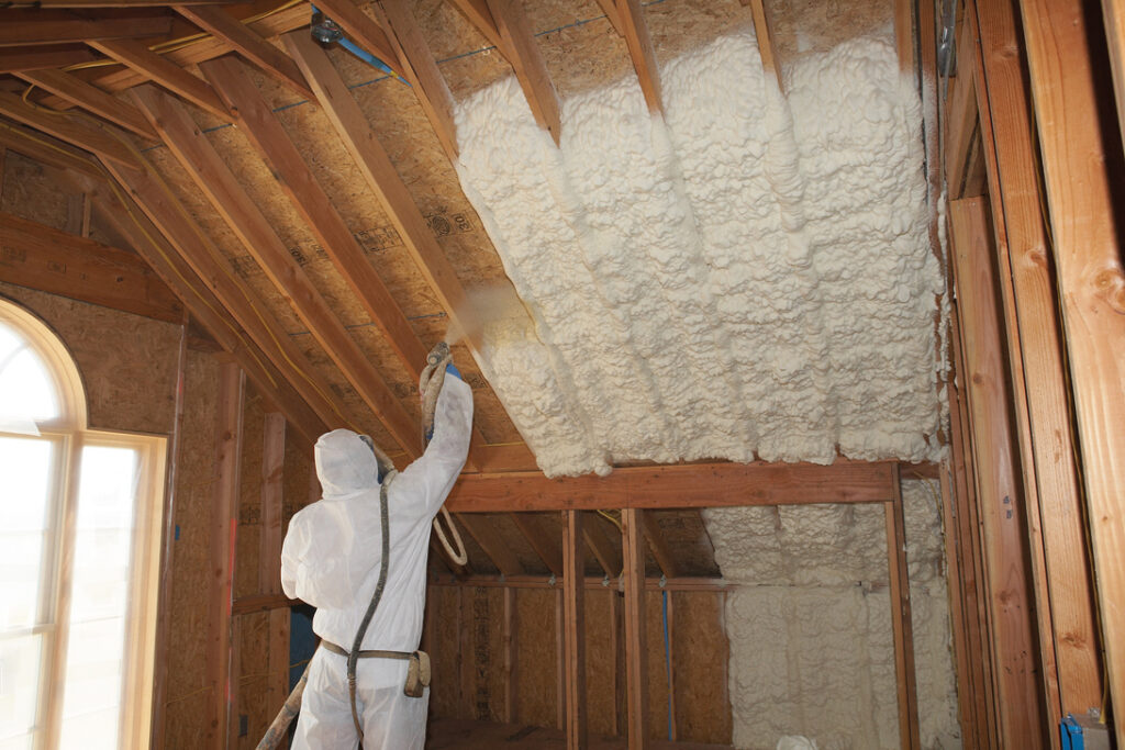 Spray foam insulation being installed in an attic ceiling by a tech in a hazmat suit